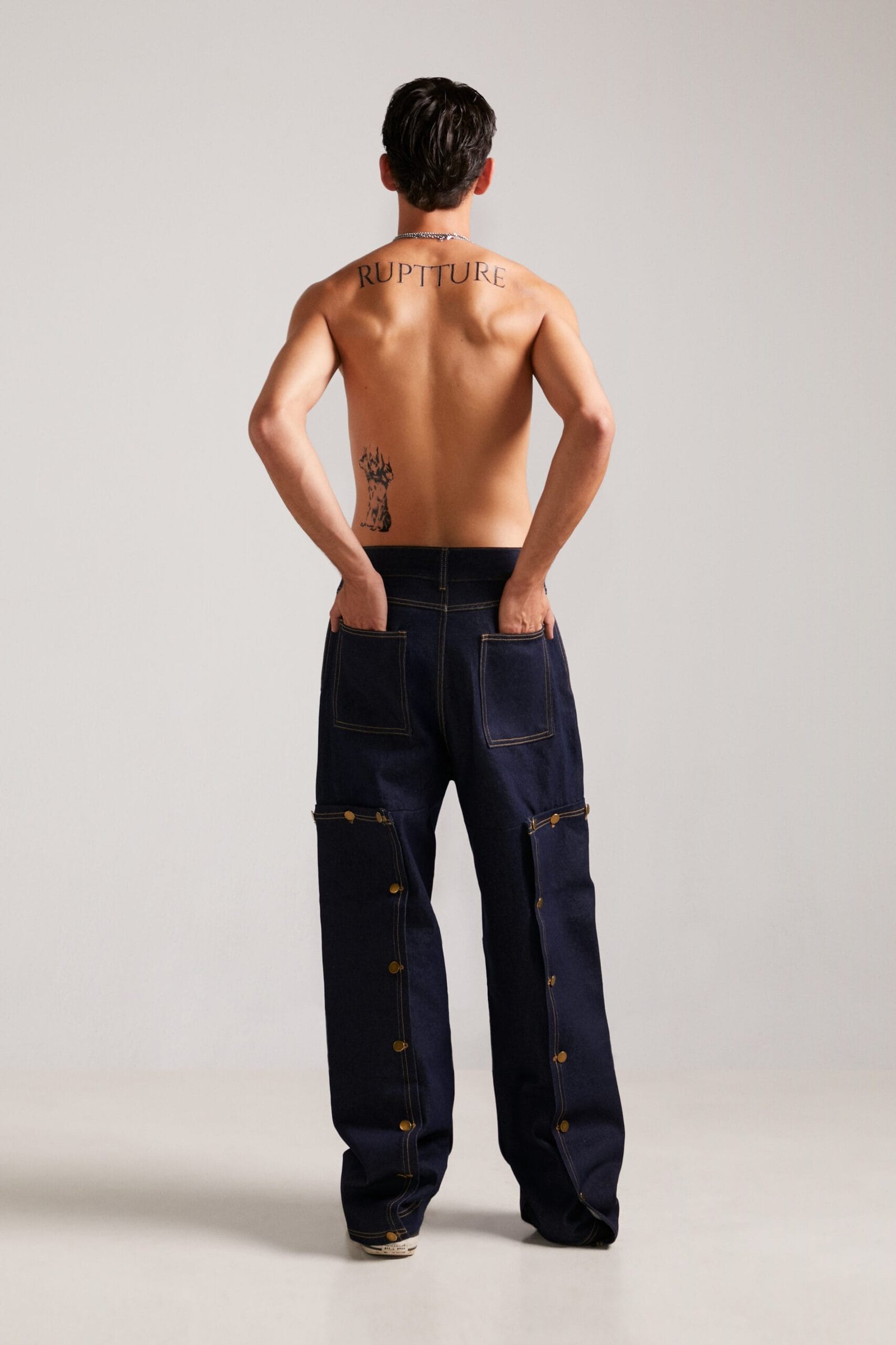 Modelo de espaldas con el tatuaje de la marca y pantalones streetwear unisex Utility Jeans denim, máxima expresión de nuestro streetwear exclusivo.