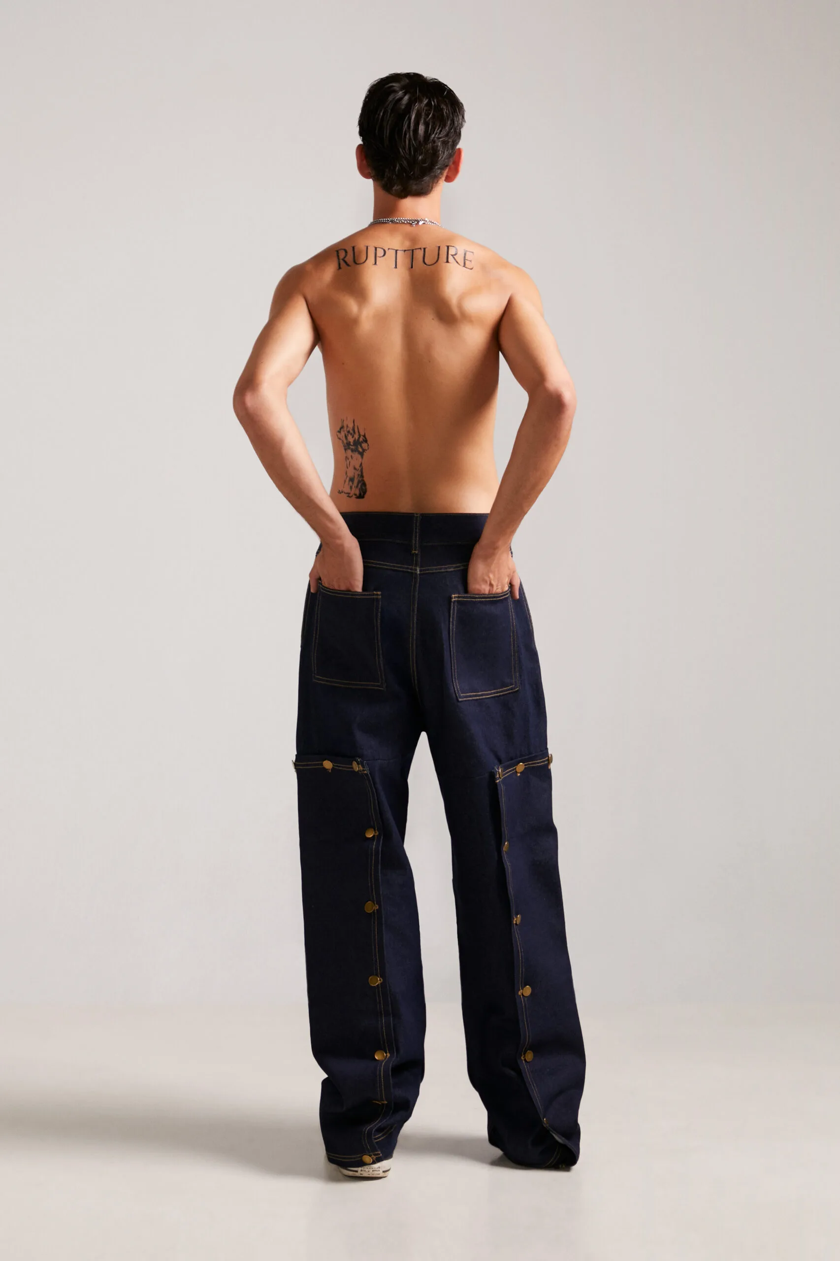 Modelo de espaldas con el tatuaje de la marca y pantalones streetwear unisex Utility Jeans denim, máxima expresión de nuestro streetwear exclusivo.