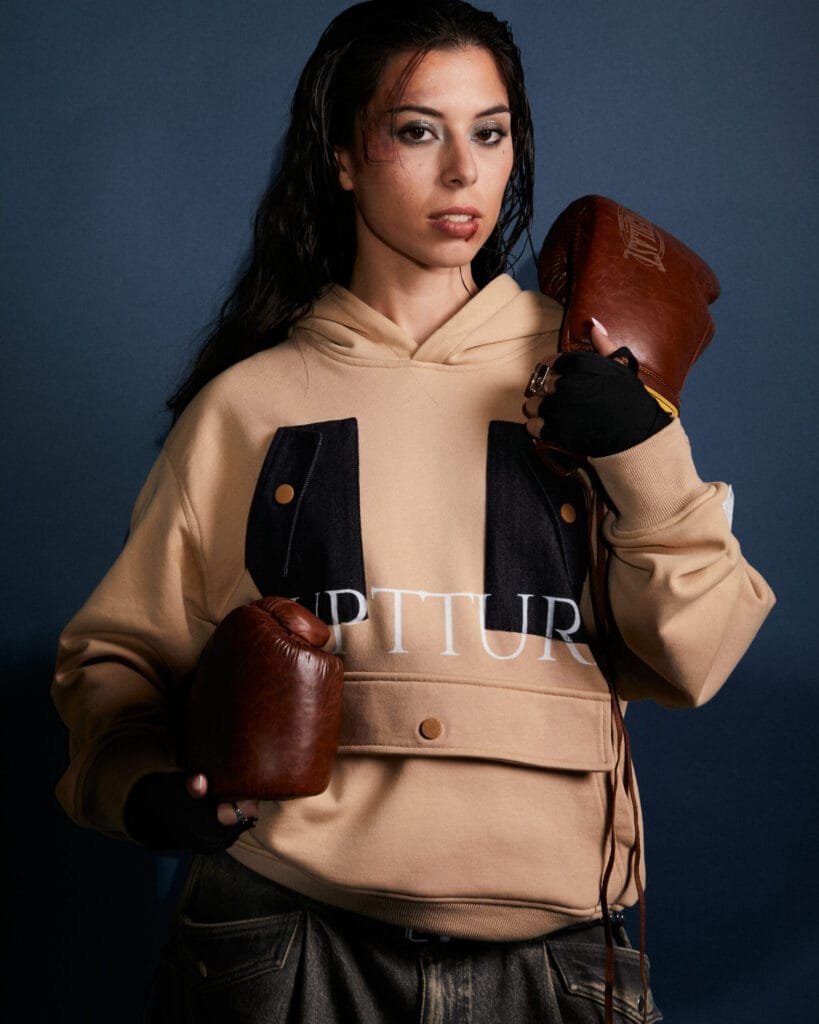 Modelo de ruptture ropa streetwear unisex con labio herido sosteniendo guantes de boxeo, luciendo la sudadera Utility Hoodie de nuestra línea de ropa streetwear de lujo.