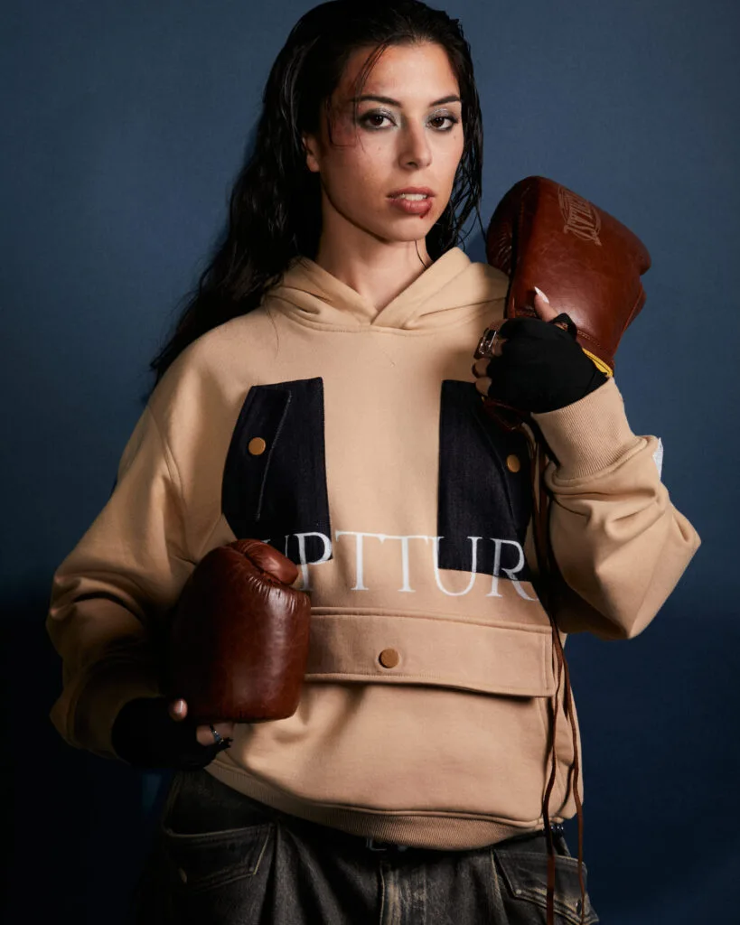 Modelo de ruptture ropa streetwear unisex con labio herido sosteniendo guantes de boxeo, luciendo la sudadera Utility Hoodie de nuestra línea de ropa streetwear de lujo.